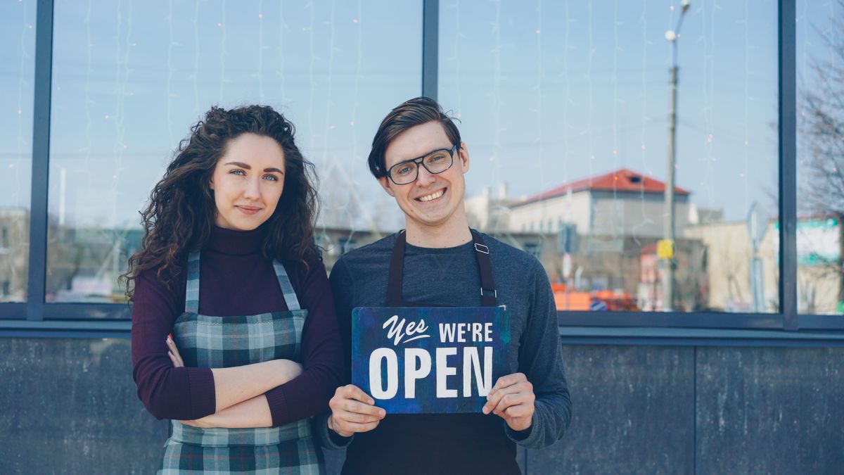 Two small-business owners holding a “Yes, we’re open” sign outside their shop